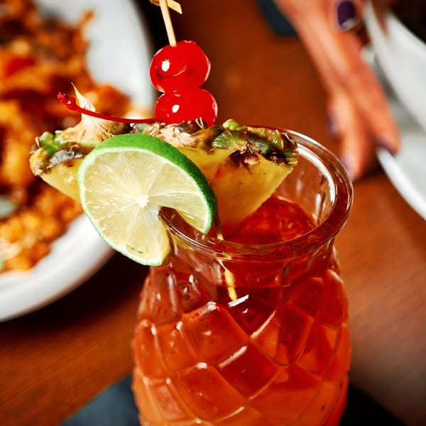 A vibrant red cocktail in a textured glass, garnished with pineapple, lime, and cherries.