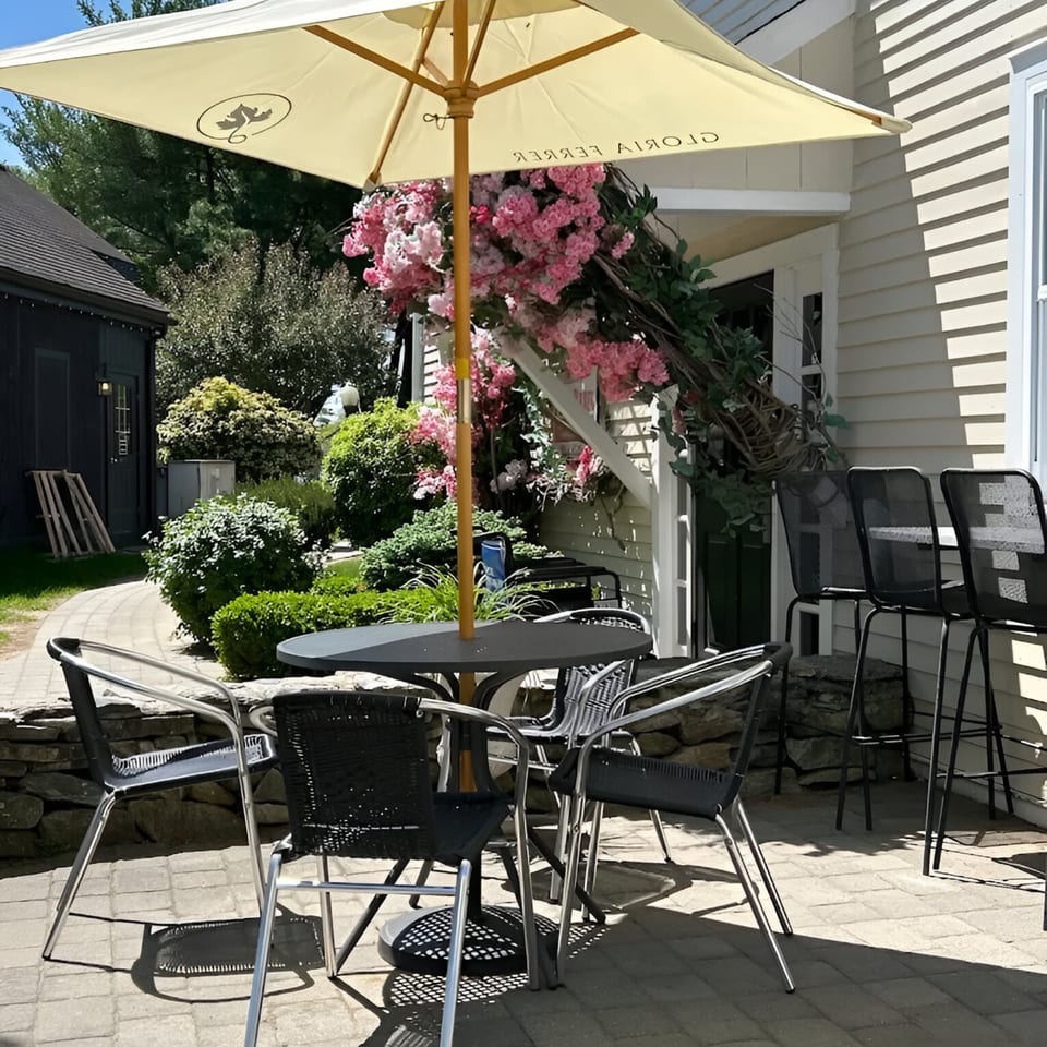 Outdoor patio with a table, chairs, umbrella, and pink flowers, next to a white building.