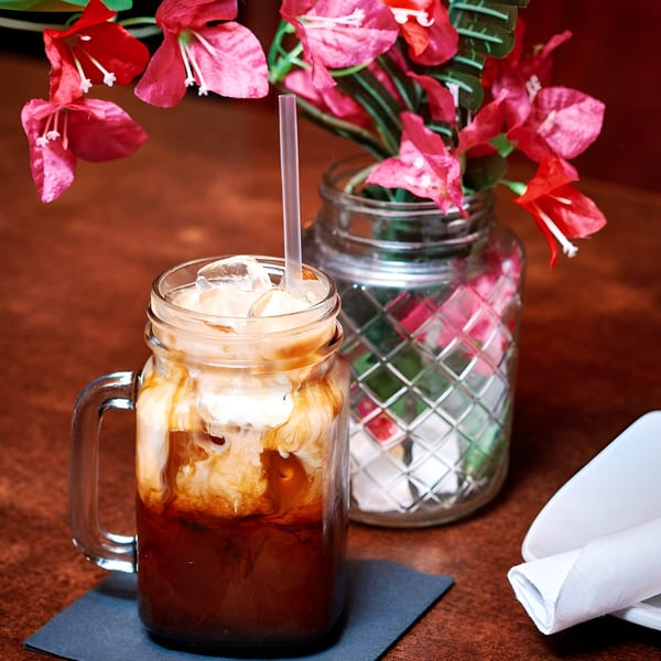 Iced Thai tea in a mason jar with a straw, next to a vase of pink flowers.
