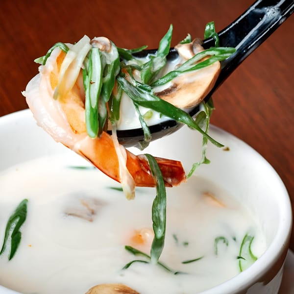 A black spoon lifts a shrimp, mushrooms, and greens from a bowl of creamy white soup.