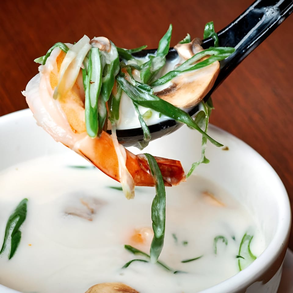 A black spoon lifts a shrimp, mushrooms, and greens from a bowl of creamy white soup.